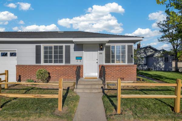 Front of the duplex: grey siding over a red brick base, a white front door between two shuttered windows, and a split-rail fence along the walk