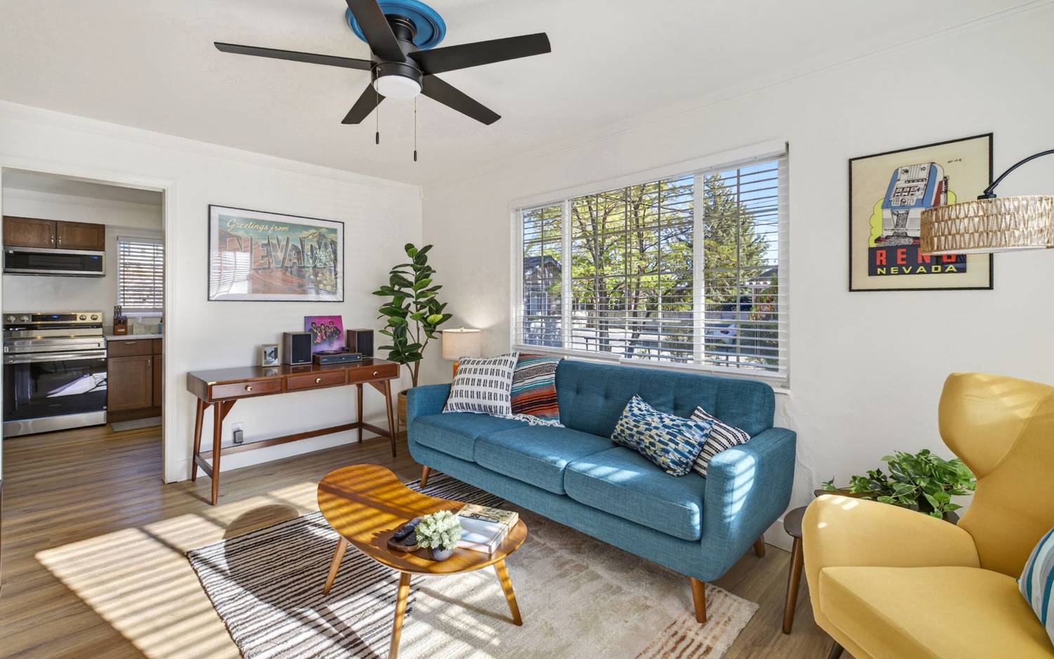 Living room with a teal tufted sofa, mustard armchair, walnut console and a low oval coffee table on an oak floor, with the kitchen visible through the doorway